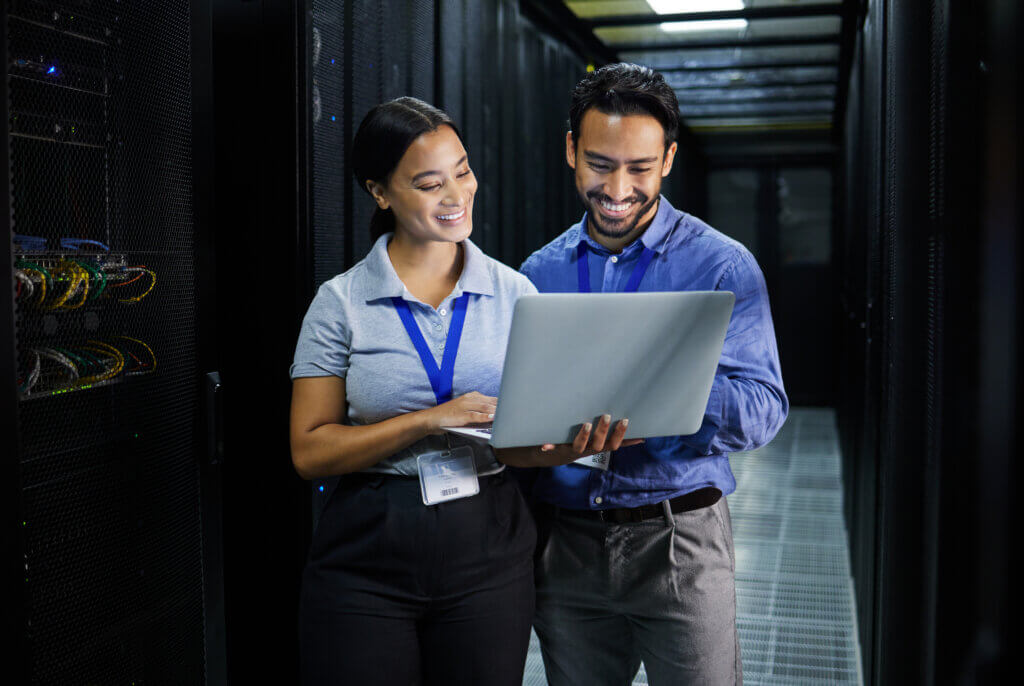 Financial Services 1 Two professionals, a man and a woman, are standing in a server room, smiling while looking at a laptop screen.