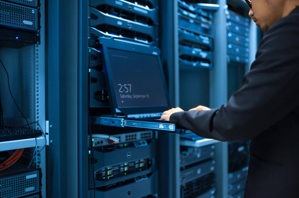 Other Industries A person in a dark outfit is working on a pull-out laptop console mounted within a server rack inside a data center, surrounded by rows of servers and networking equipment.