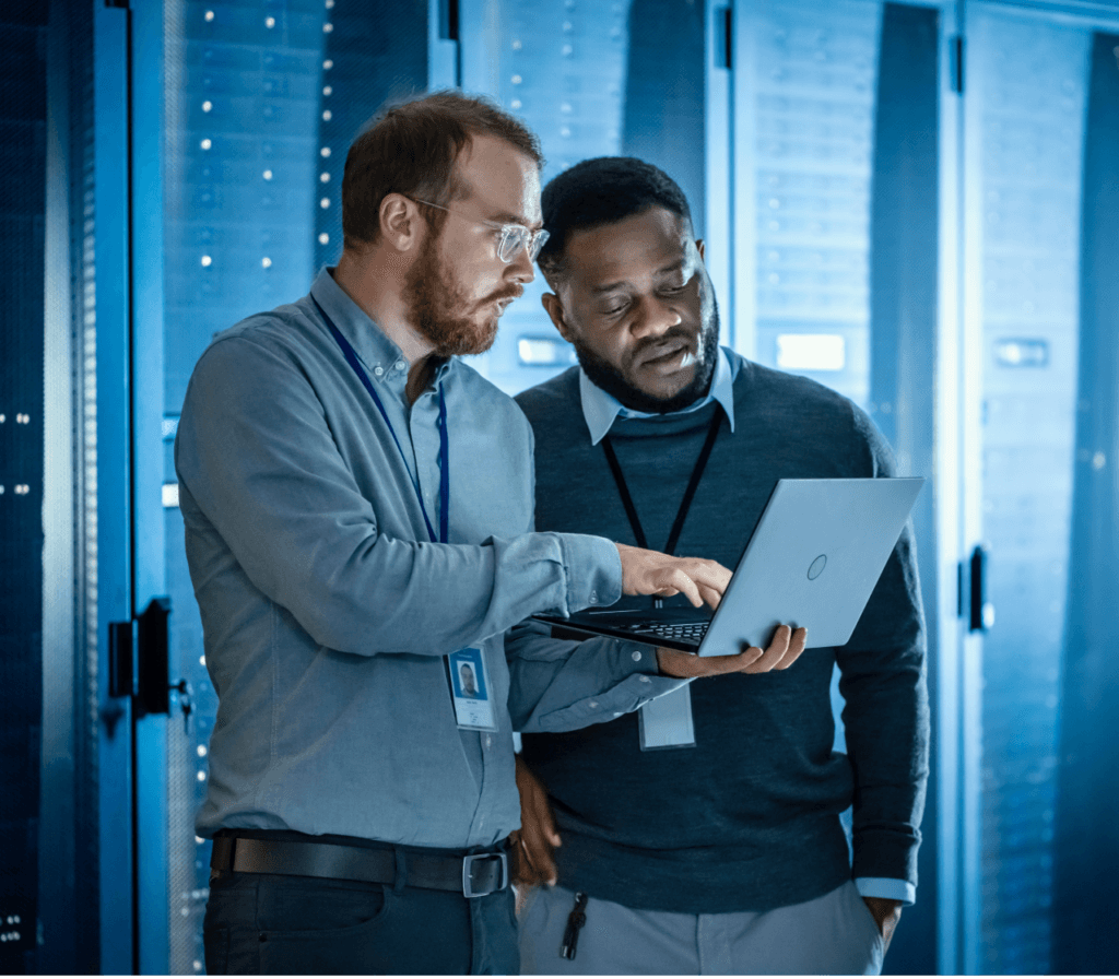 Two IT professionals in a server room analyzing data on a laptop, surrounded by rows of illuminated server racks.