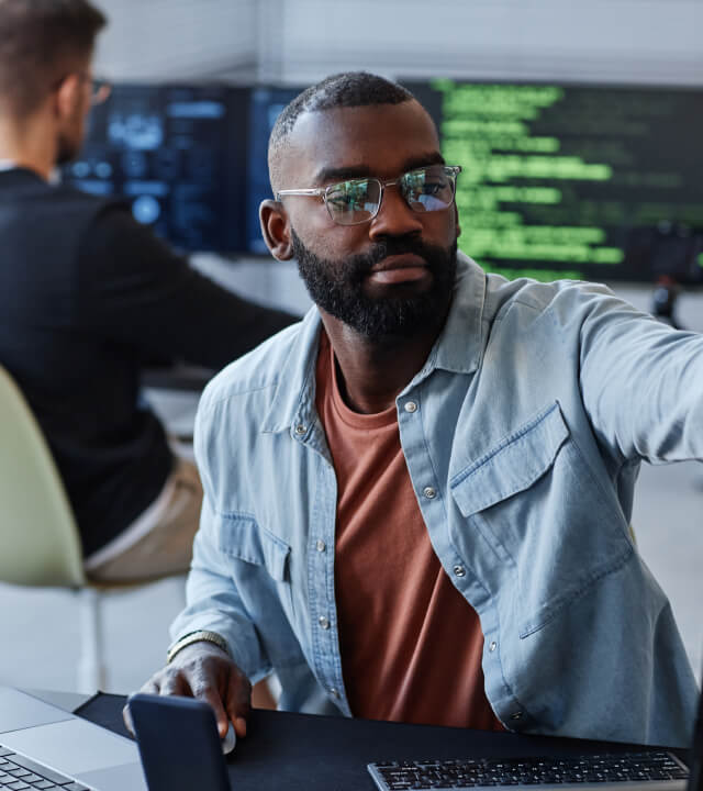 Careers A man with a beard and glasses, wearing a denim shirt over a rust-colored t-shirt, sits at a desk with a smartphone and keyboard, while pointing to something off-screen