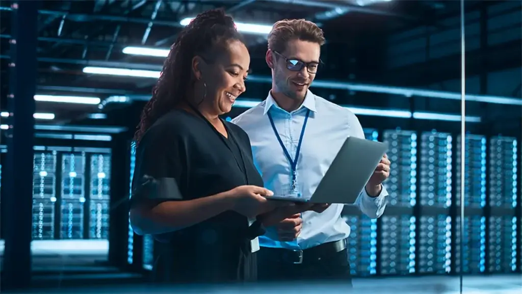 A woman and a man are smiling and looking at a laptop in a server room, surrounded by rows of server racks with blue lighting. A woman and a man are smiling and looking at a laptop in a server room, surrounded by rows of server racks with blue lighting.